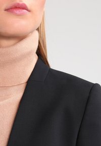 Svart kavaj med en strukturerad krage, kombinerad med en ljusbeige polotröja. Mjuk textur och en figursydd silhuett.