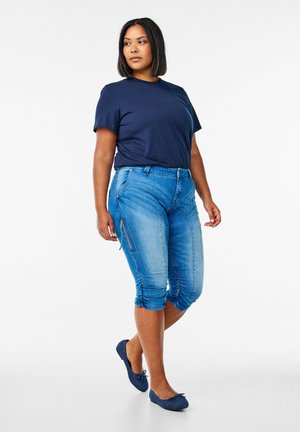 Camiseta de manga corta de color marino combinada con pantalones capri de mezclilla azul claro, que cuentan con cremalleras laterales y detalles fruncidos en el dobladillo. Se lleva con zapatos planos azules.