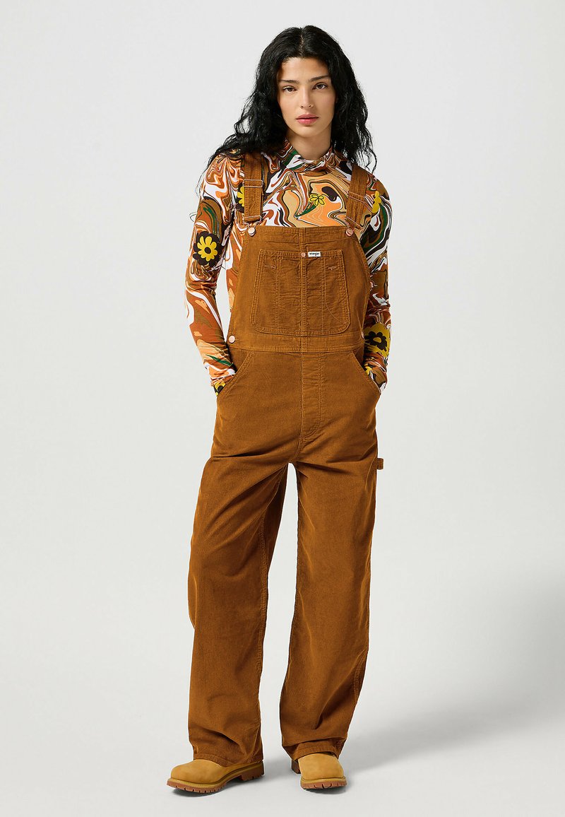 Brown corduroy overalls with adjustable straps, side pockets, and contrasting buttons, paired with a long-sleeve patterned top and boots.