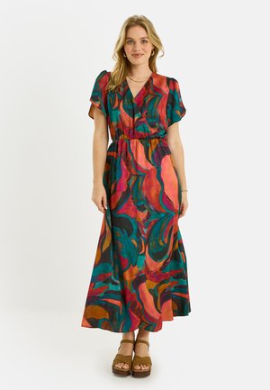Mujer de pie, lleva un vestido largo multicolor con patrón abstracto, mangas cortas y sandalias marrones con plataforma, sonriendo ligeramente.