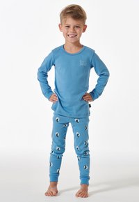 Haut de pyjama bleu à manches longues avec un design d'ours, associé à un pantalon bleu clair avec des motifs de lune et de petits points rouges. Matière en coton doux.