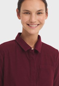 Camisa de pana burdeos con cierre de botones en la parte delantera, cuello y bolsillo en el pecho. Textura suave con costuras visibles y detalles de canalé.