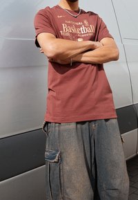 Burgundy T-paita, jossa on kultainen NBA-teksti ja koripallografiikka, yhdistettynä sinisiin cargo-shortseihin, joissa on taskut ja haalistunut rakenne.