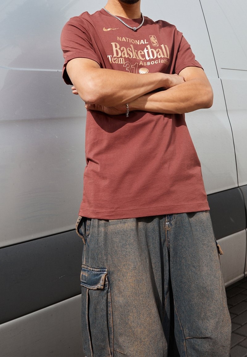 Burgundy T-paita, jossa on kultainen NBA-teksti ja koripallografiikka, yhdistettynä sinisiin cargo-shortseihin, joissa on taskut ja haalistunut rakenne.