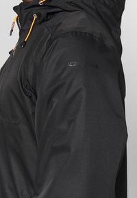 Sort vandtæt jakke med et struktureret design, justerbare orange snore og et logo på venstre skulder.