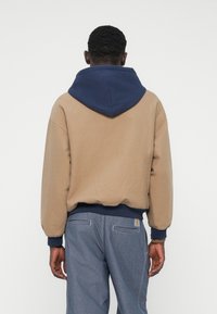 Hnedá a tmavomodrá mikina s kapucňou s voľným strihom, rebrovanými manžetami a lemem. Hladká textúra s dvojvrstvovým dizajnom kapucne.