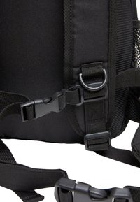 Melnas auduma mugursoma ar regulējamām siksnām, plastmasas sprādzēm un sieta kabatu, uzsverot izturīgu konstrukciju un funkcionalitāti.