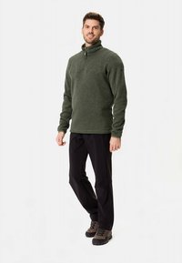 Maglione verde in pile con mezza cerniera, tessuto strutturato e tasca sul petto. Abbinato a pantaloni neri e scarpe da trekking marroni.