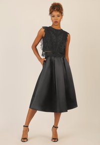 Top corto in pizzo nero con bordi ondulati abbinato a una gonna nera a linea A in satin. Presenta tasche; il modello indossa dei sandali neri con fasce.
