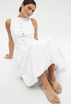 White sleeveless dress with a high neckline and front button closure, paired with beige cross-strap sandals. Smooth texture, flowing silhouette.