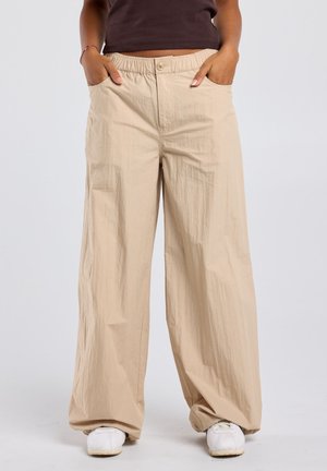 Persoon draagt beige broek met wijde pijpen en elastische tailleband met knoop, handen in de zakken, gecombineerd met witte sneakers.