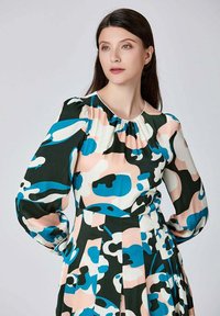 Femme aux longs cheveux noirs portant une robe à manches longues avec des formes abstraites bleues, noires, blanches et pêche sur un fond uni.