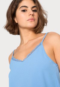 Camisola azul-clara com delicadas rendas ao longo da gola em V e alças finas ajustáveis. Tecido suave e liso com um corte ajustado.