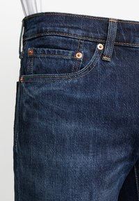 Tummansinisiä farkkuhousuja, joissa on suora leikkaus, kupariset niitit, näkyvät ompeleet, viisi taskua ja sileä pinta.