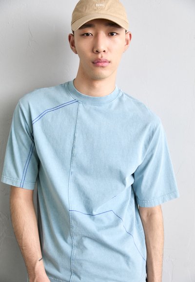 T-shirt en coton bleu clair avec un motif de couture irrégulier en bleu foncé, à manches courtes et col rond.