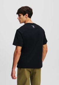 Juodas medvilnės marškinėliai trumpomis rankovėmis, apvaliu iškirpimu ir mažai grafika ant nugaros. Modelis vilki juos su alyvuogių žalios spalvos kelnėmis.