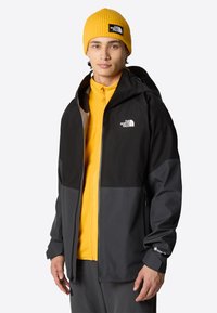 Czarna i szara kurtka z kapturem z widocznymi zamkami, noszona na żółtej polarowej bluzie. Na piersi i czapce znajduje się logo The North Face.