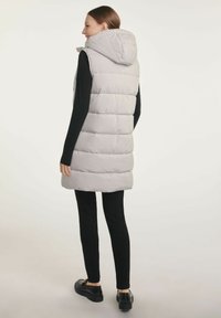 Jasnoszara kamizelka puchowa z kapturem, z poziomymi przeszyciami i dłuższym fasonem, w połączeniu z czarnymi dopasowanymi spodniami i czarnymi butami.