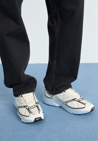 Bílé tenisky Nike s černými prvky, mají síťovaný svršek, zaoblenou špičku a tlustou podešev. Nositelná s černými džínovými kalhotami na modrém pozadí.