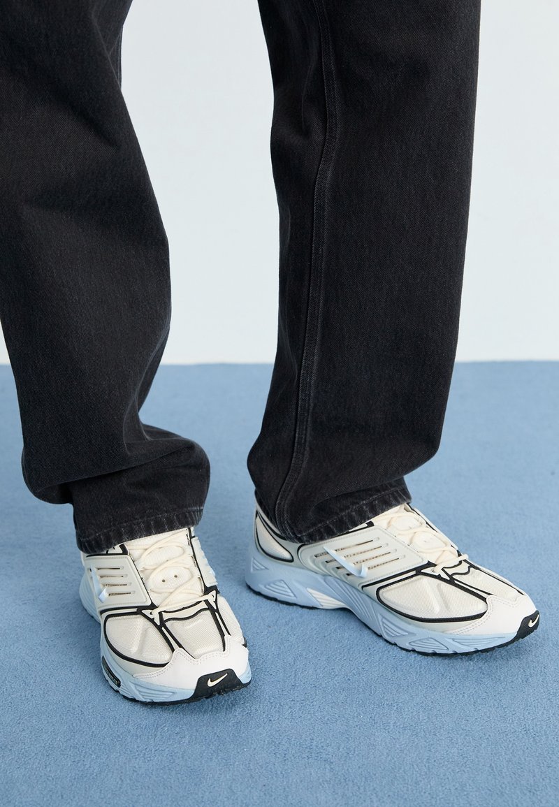 Vita Nike-sneakers med svarta detaljer, med meshtopp, rundad tå och tjock sula. Bärs med svarta denimbyxor mot en blå bakgrund.