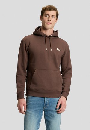Hombre con sudadera marrón con capucha y el logotipo "Lee" en el pecho y vaqueros azul claro, de pie sobre un fondo liso gris claro.