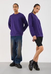 Twee modellen met gevlochten haar dragen bijpassende paarse sweatshirts; de een combineert het met losse jeans en zwarte sneakers, de ander met zwarte fietsbroek en laarzen.