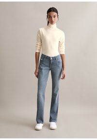 Lichtbeige coltrui, aansluitende mouwen; blauwe spijkerbroek met rechte pijpen, 5-pocket ontwerp; witte sneakers; neutrale achtergrond.