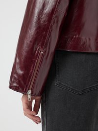 Burgundy leather jacket with a glossy finish, featuring a zipper at the cuff and a relaxed fit. Gray denim background with pocket detail.