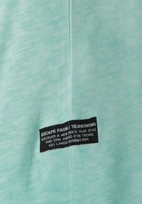 T-shirt verde menta con tessuto testurizzato. Presenta un'etichetta nera con testo bianco, che fornisce informazioni sul marchio e un tagline.