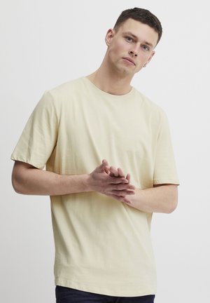 Pale yellow short-sleeve t-shirt made of soft fabric with a round neckline, smooth texture, and a relaxed fit. Hands clasped in front.