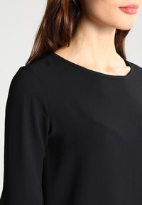 Maglietta nera a maniche lunghe con scollatura tonda e superficie testurizzata. Il tessuto sembra leggero e liscio, con dettagli sottili alle maniche.