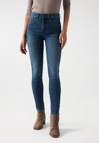 Vaqueros ajustados azul oscuro con una cintura alta, que cuentan con bolsillos frontales y una sutil textura desgastada. Combinados con botas de ante grises hasta el tobillo.
