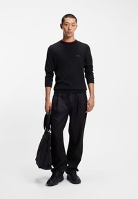 Maglione di lana nero con collo a girocollo rosso, abbinato a pantaloni neri rilassati. Il modello tiene una borsa nera e indossa sneakers nere.