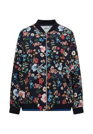 Frau mit beiger Cap und marineblauer Bomberjacke mit mehrfarbigen Blumen, lächelnd vor neutralem Hintergrund.