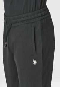 Pantaloni sportivi neri realizzati in tessuto morbido, con elastiche in vita, tasche laterali, cordoncino e un piccolo dettaglio in metallo sul gamba.