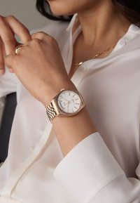 Montre en métal doré avec un cadran blanc, des index argentés et un bracelet à maillons, portée sur un poignet partiellement couvert par une chemise blanche.