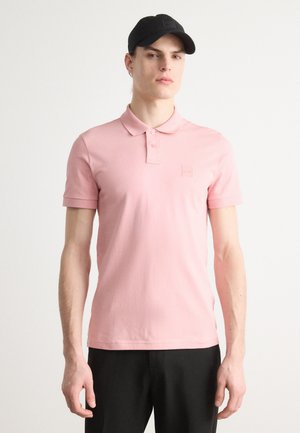 Rosa polo med krage, korte ermer og et lite logo-lapp på venstre bryst. Brukt med svarte bukser og en svart caps.