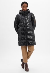 Schwarze Daunenjacke mit glänzender Textur, Kapuze, Kordelzug und Futter im Leopardenmuster. Kombiniert mit hellbraunen Hosen und braunen Knöchelstiefeln.