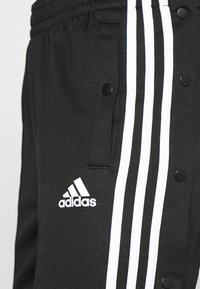 Svarta träningsbyxor med vitt Adidas-logotyp och tre vertikala ränder. Har tryckknappsfickor och en texturerad tyg.