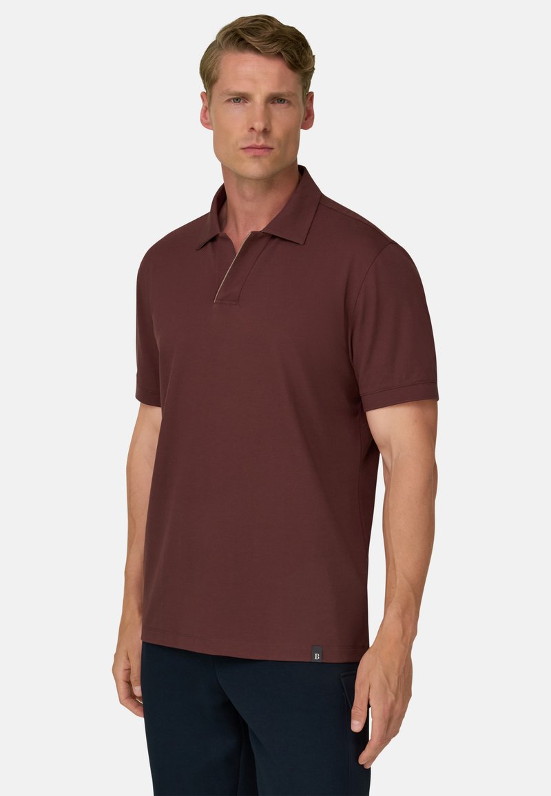 Homme portant un polo bordeaux à manches courtes et un pantalon foncé, debout devant un fond clair uni.