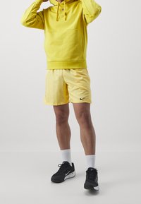 Sweat à capuche jaune avec une poche kangourou, assorti à un short jaune arborant un logo Nike noir. Chaussures de sport noires et chaussettes blanches.