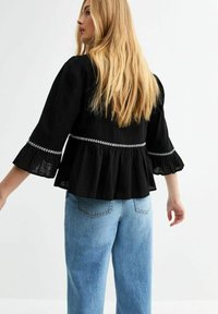 Blusa nera a peplo con maniche a campana, con ricami bianchi in vita, abbinata a jeans azzurri a vita alta.