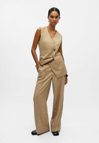 Gilet beige ajusté avec un décolleté en V profond et deux boutons, associé à un pantalon à jambes larges. Texture lisse avec un motif de tissage subtil et des poches.