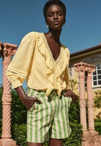 Blusa gialla a volant con bordi all'uncinetto, abbinata a pantaloni corti verdi e crema a righe con dettagli in bottone e tasche laterali.