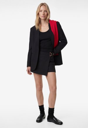 Mujer vistiendo blazer negra sobre top negro y minifalda con hebilla, calcetines negros y mocasines, sosteniendo una chaqueta roja sobre el hombro.