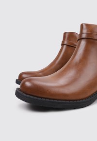 Botines de cuero marrón con acabado liso, punta redonda y suela de goma. Presentan una discreta correa en el tobillo como detalle adicional.