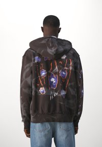 Sudadera con capucha en camuflaje oscuro con un vibrante diseño floral azul, presenta un dobladillo y puños desgatados. Modelo vistiendo con jeans de un azul claro.