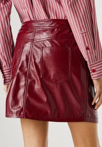 Mini-jupe en simili cuir bordeaux avec une texture brillante. Dotée de poches arrière et d'une ceinture visible embossée avec le logo de la marque.