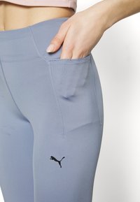 Jasnoniebieskie legginsy sportowe z elastycznego materiału, z boczną kieszenią i małym czarnym logo Pumy na udzie.
