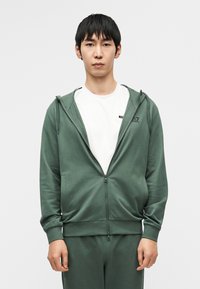 Roheline lukkudega kapuutsiga pusa koos kängurutaskuga, kantud valge särke peal. Pehme kangas sileda tekstuuriga, millel on soonilised varrukasuu ja taljeäär.